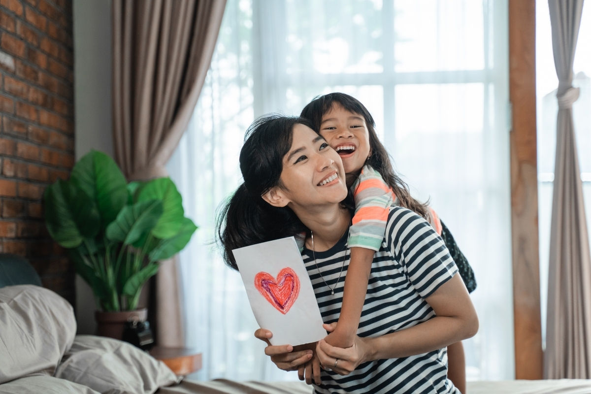 A young girl hugging her mom who has a card in hand for Mother's Day
