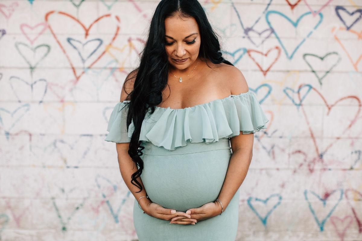 A fancily dressed pregnant woman posing with her belly in front of a wall covered in graffiti hearts.