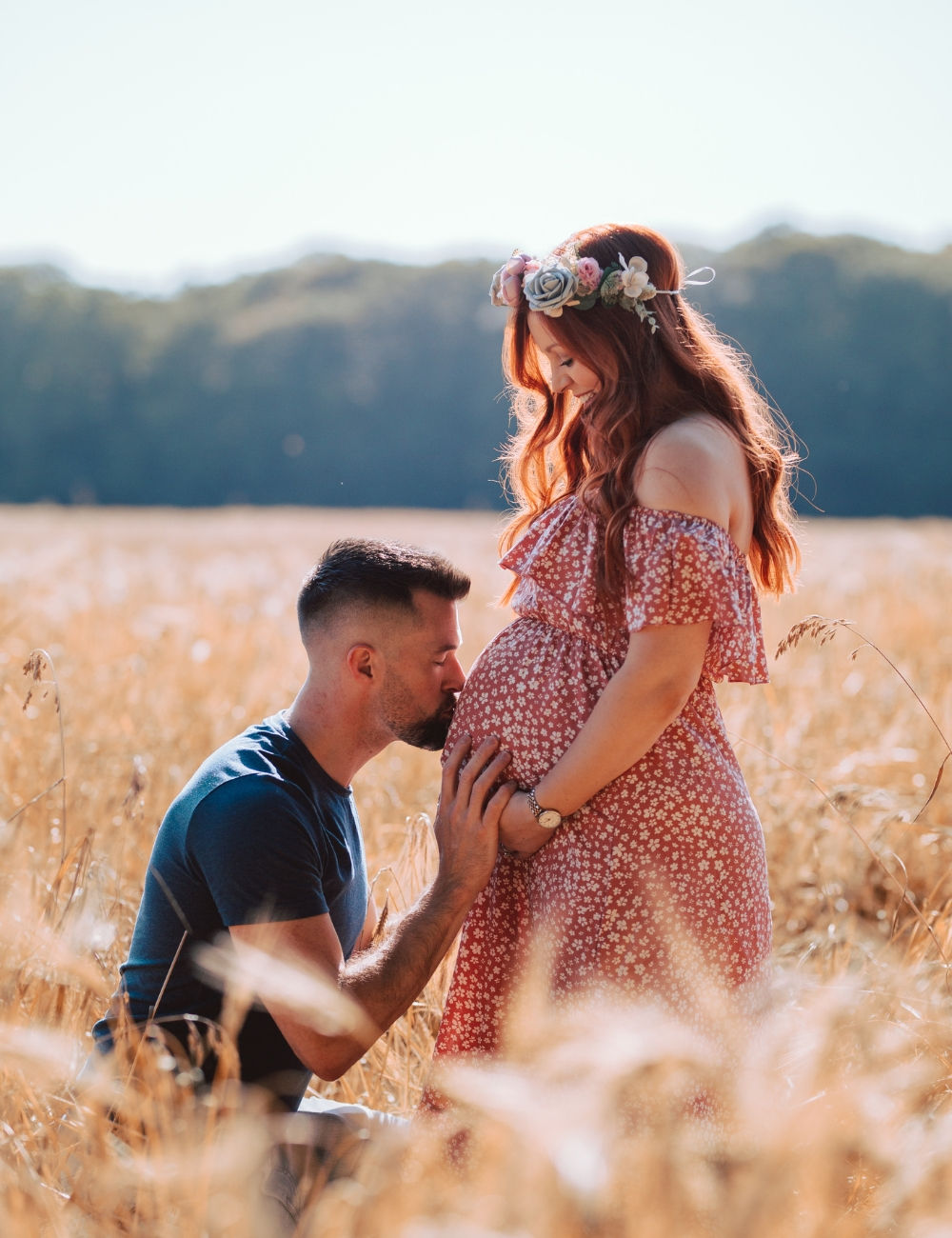 A man kneeling and kissing his pregnant partners belly in a wheat field