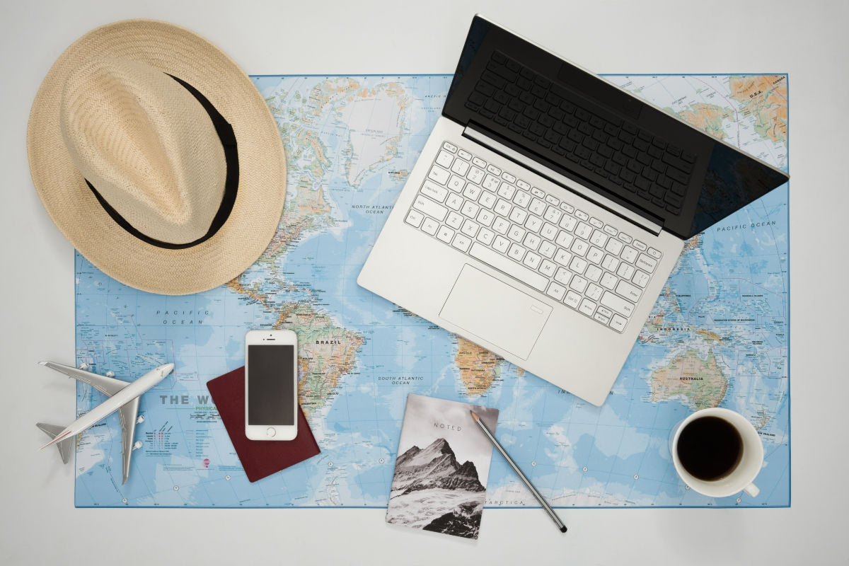 Laptop, map, phone, passport, hat, airplane model, coffee on table suggest travel planning mood. Notebook labeled "NOTED".