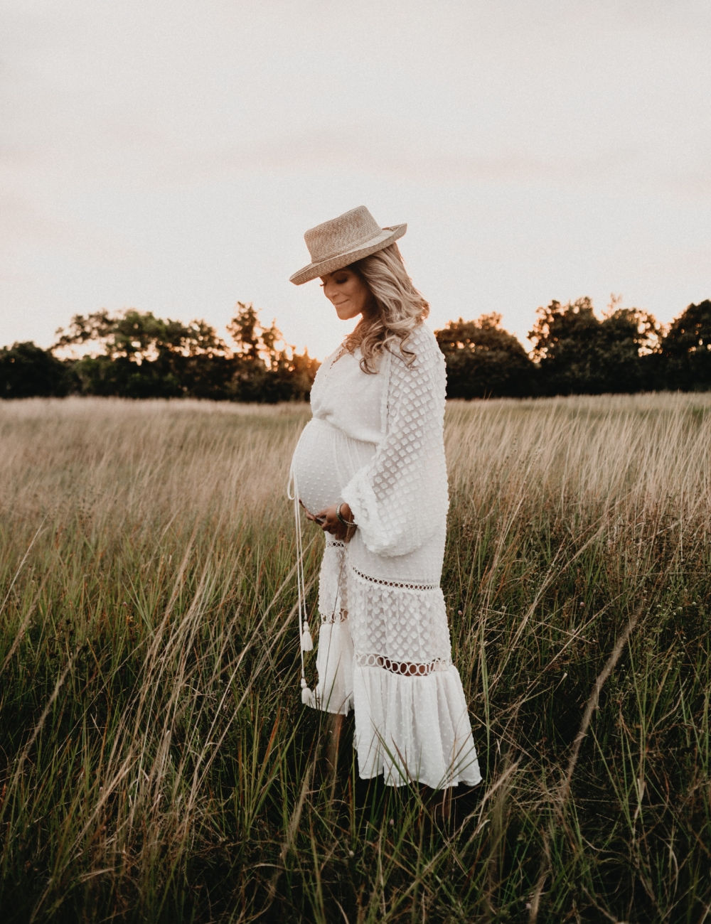 A pregnant woman in a bohemian white maxi dress holding her belly in a field at sunset