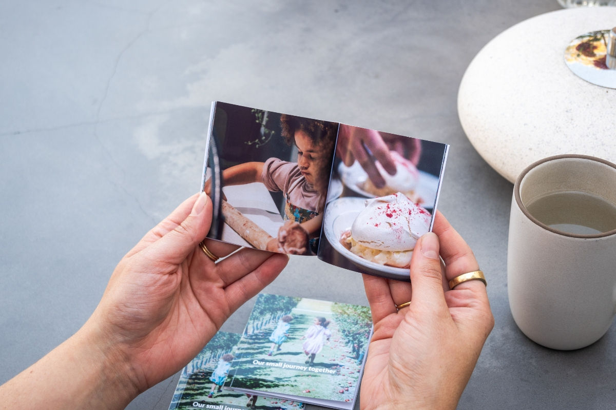 Hands hold a small photo book showing a child rolling dough and a dessert. Background: cup, coaster, gray surface.