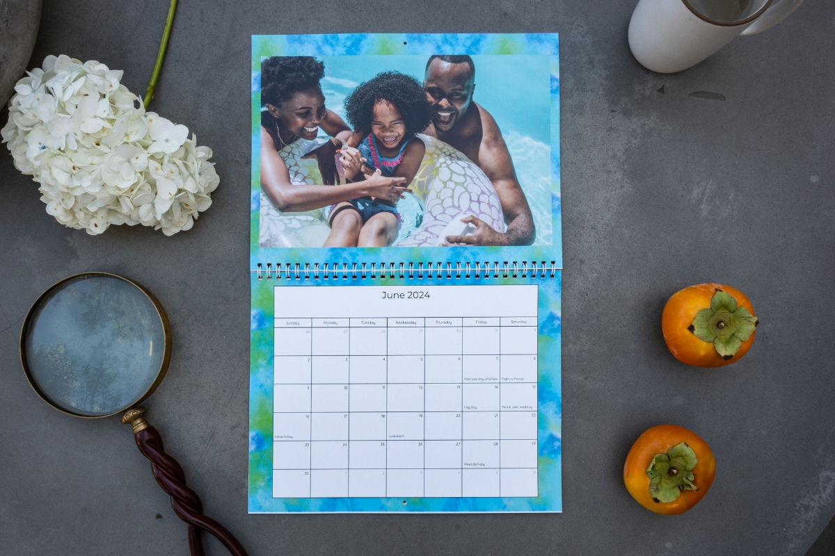 Calendar open to June 2024, with a photo of a joyful family in a pool. Nearby are hydrangeas, a magnifying glass, and two persimmons.