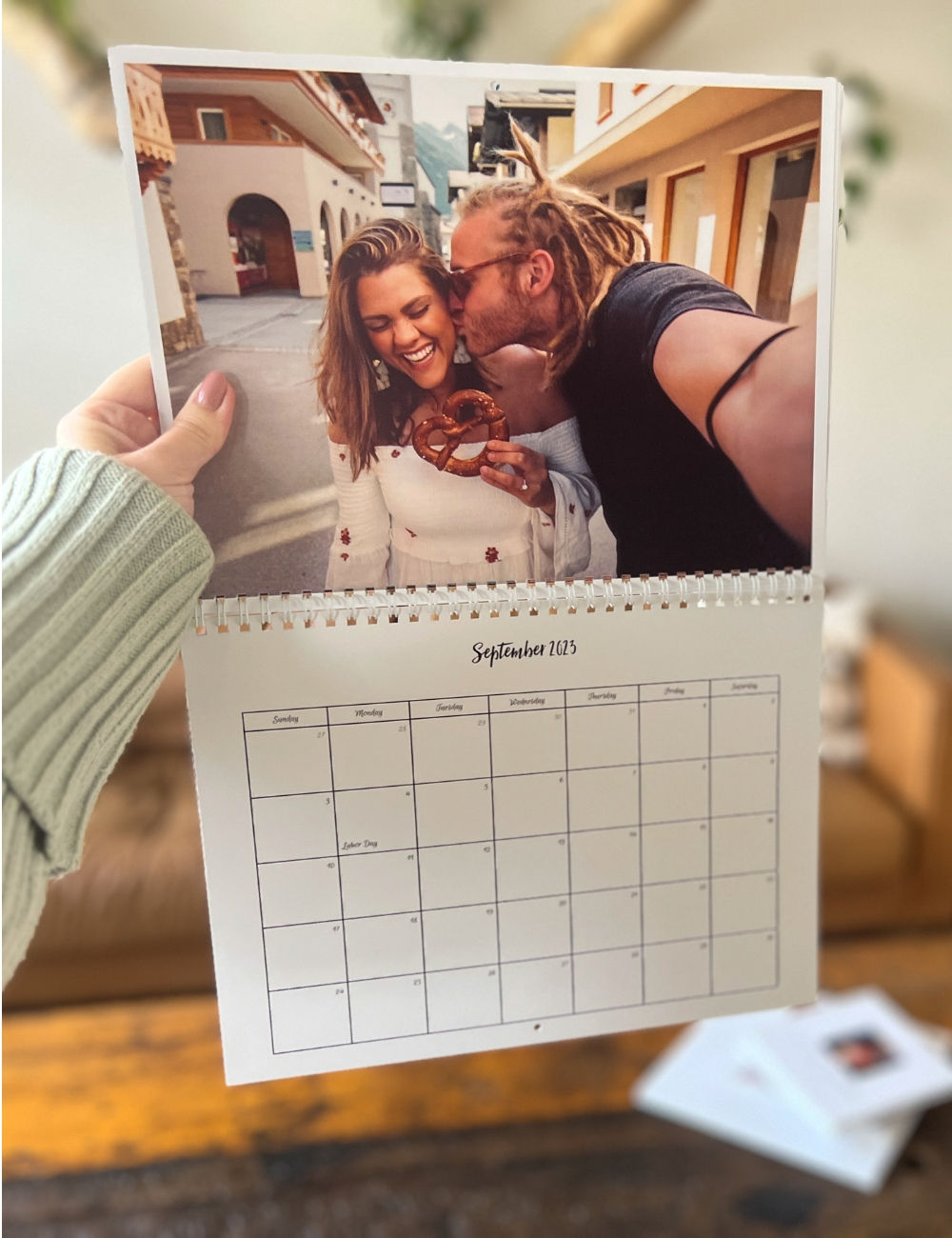 a hand holding a calendar up with a couple in a quaint street — the women is holding a pretzel and the man is taking a selfie as he kisses her on the cheek