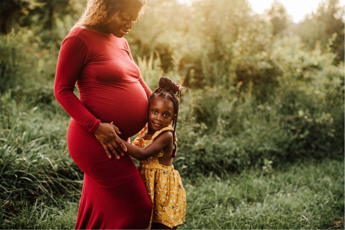 A fancily dressed pregnant woman posing with her toddler with a lush green background