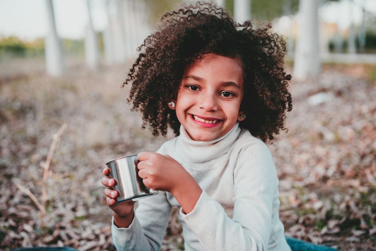 A kid smiling at the camera with a warm cup of cider in her hands