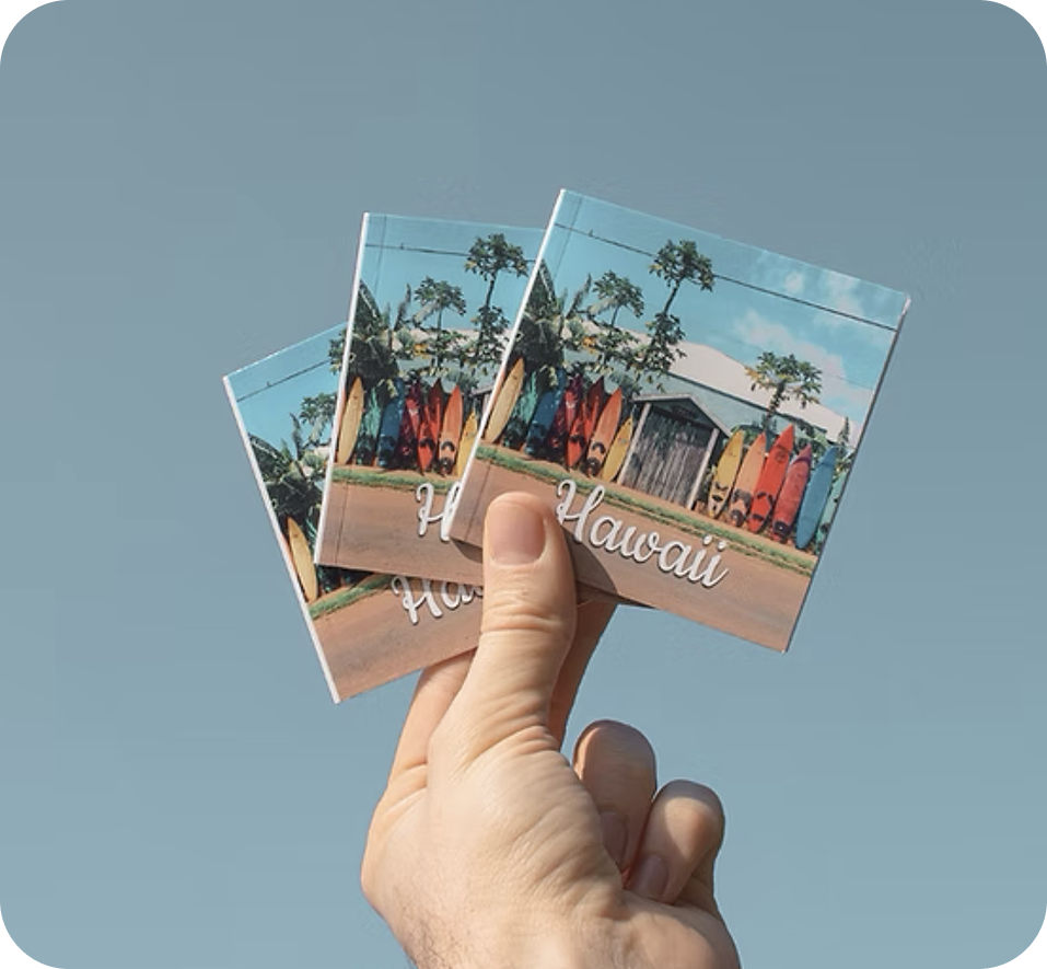 A hand holding up three mini photo books with a Hawaii cover against a blue sky
