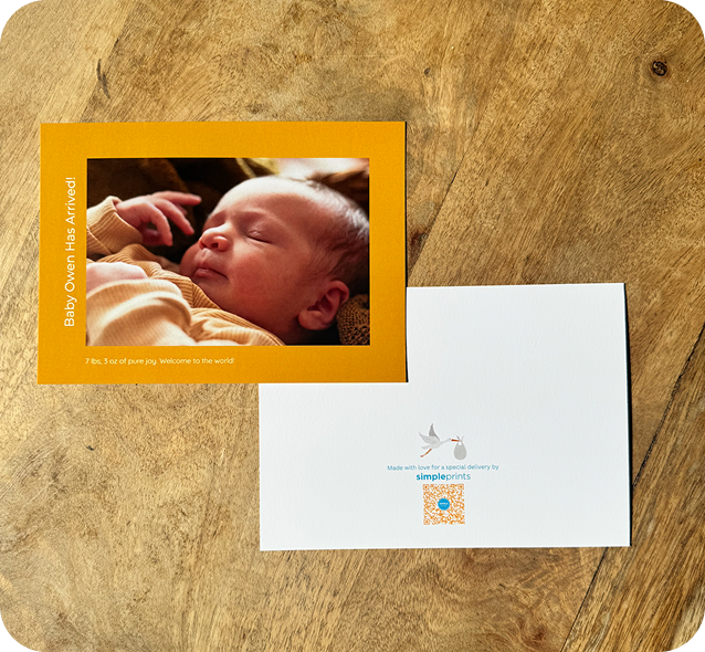 A birth announcement photo card of a sleeping newborn beside its printed back, on a wooden table
