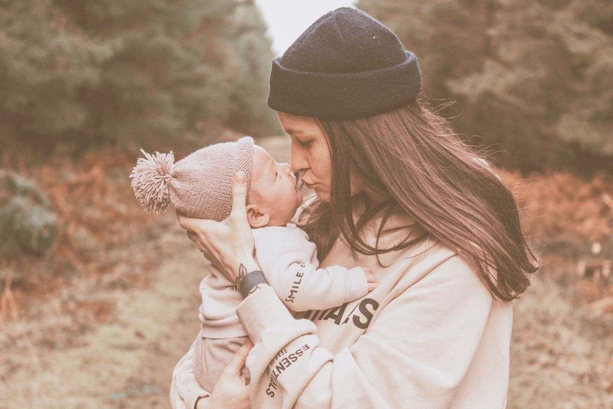 Woman in a beanie kisses a baby wearing a pom-pom hat in a forest. Warm tones, cozy attire, and lettering on her sleeve visible. Personalized Mother's Day Gift ideas to make her feel extra special on the day!