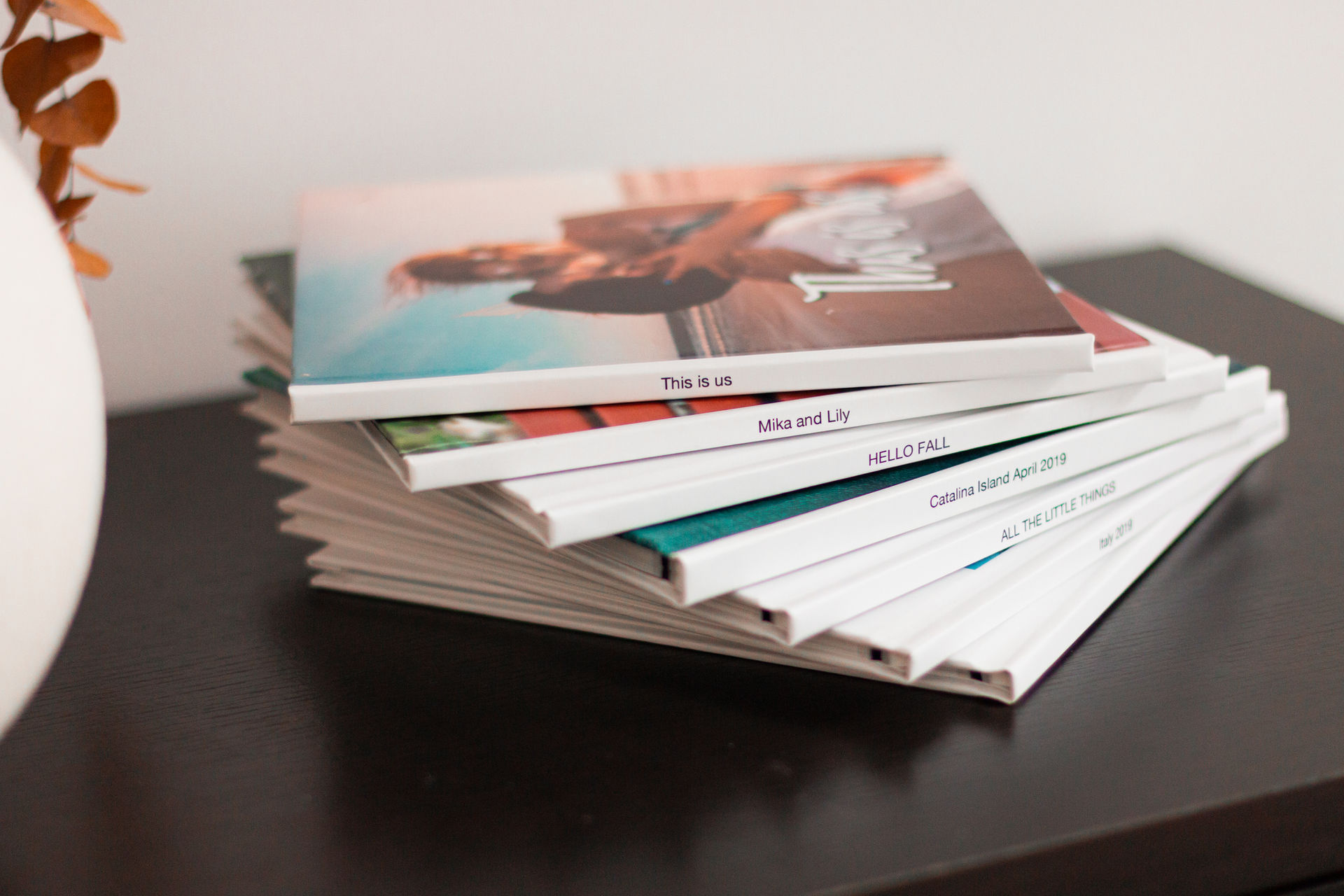 A stack of hardcover photo books on a dark shelf, their spines printed with titles such as This is us and HELLO FALL